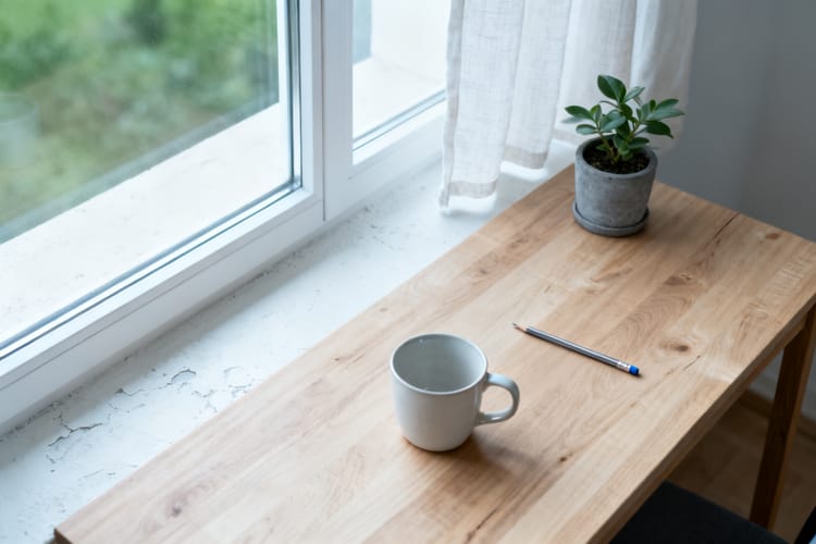 Leerer heller Schreibtisch am Fenster mit einem Stift und einer Tasse, weiches Vormittagslicht